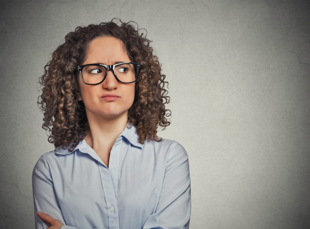 Displeased suspicious young woman with glasses looking sideways isolated on grey wall background. Negative face expression emotion perception